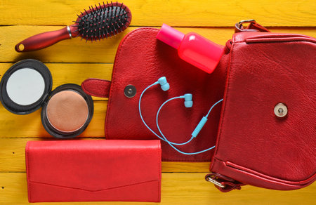 What's in the women's bag? A red bag, leather purse, headphones, mirror, comb, perfume bottle on a yellow wooden table. Top view.の写真素材
