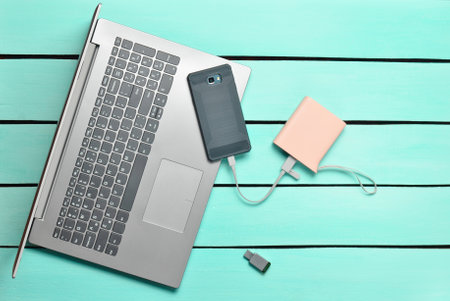 Laptop, smartphone, power bank, USB flash drive on a blue wooden table. Modern digital devices and...
