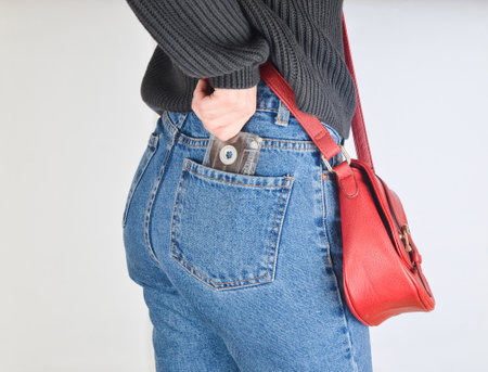 A girl in a mimes fit jeans, a sweater and with a red bag on the edge pulls out a retro audio cassette from her pocket on a white background. Side view.の写真素材