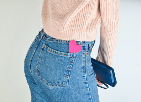 Decorative heart in the back pockets of jeans of a slim girl who holds a purse in her hands on a...