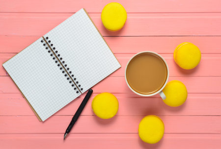 A notebook with a pen, a cup of coffee and yellow macaroons on a pink wooden table. Top view. Flat lay.の写真素材
