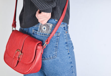 A girl in a mimes fit jeans, a sweater and with a red bag on the edge pulls out a retro audio cassette from her pocket on a white background. Side view.の写真素材