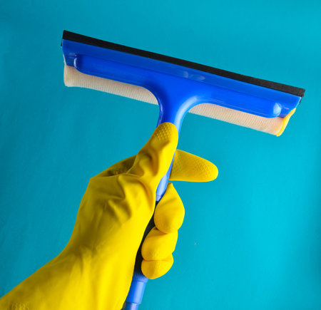 A hand with yellow rubber gloves holds a window mop on a blue background. Cleaning conceptの写真素材