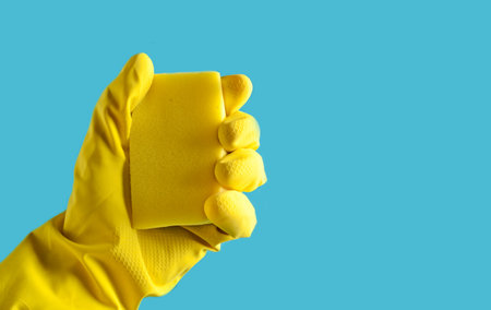 Hand with yellow rubber glove holds a sponge on a blue background, copy space