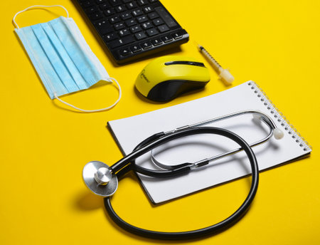 Workplace of a modern doctor. Keyboard, wireless mouse, notebook, stethoscope, syringe on yellow background.の写真素材