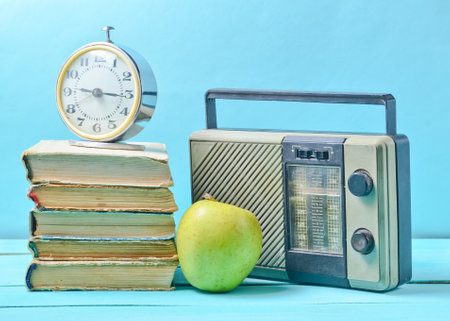 Alarm clock on stack of old books, radio receiver, apple on a blue background. Retro still life