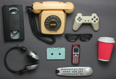 Retro objects on gray background. Rotary telephone, audio cassette, video cassette, gamepad, 3d glasses, tv remote, headphones, push-button phone. Analog media technology of the past. Flat lay.の写真素材