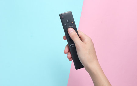 Female hand holds the TV remote on a pink blue pastel background. Top View