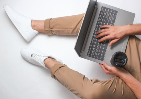 Fragment of male legs in beige pants and white sneakers sitting on white. Man using modern laptop. Online worker, freelancing, work at home, man working. top view on white backgroundの写真素材