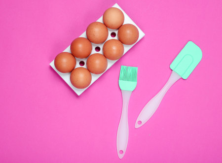 Silicone kitchen tools (spatula, brush), tray of eggs on pink background. Cooking concept, Top view,...