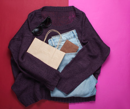 Flat lay style burgundy knitted sweater, trendy blue jeans, paper bag and other accessories on red...