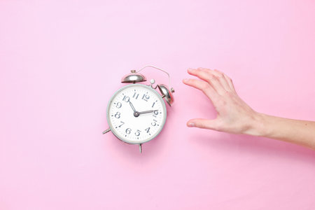 Female hand takes alarm clock on pink background. Top view. Time to wake up. Minimalismの写真素材