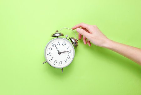 Female hand takes alarm clock on green background. Top view. Time to wake up. Minimalism