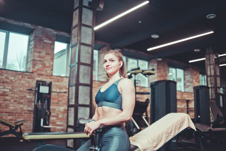 Training pause, sporty young woman in sportswear resting on bench holding dumbbells in her hands and listens to music in headphones at gymの写真素材