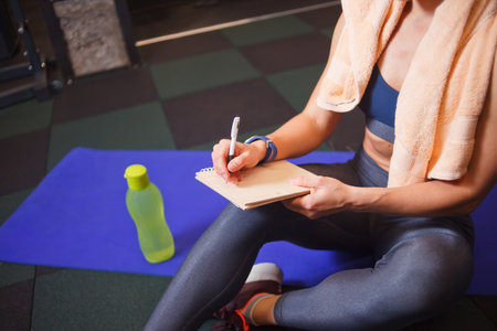 Sporty woman sits on a training mat and writes down future training plans for achieving great results in gymの写真素材