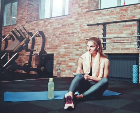 Cheerful sporty woman sits on a training mat and writes down future training plans for achieving great results in gymの写真素材