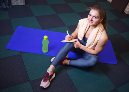 Cheerful sporty woman sits and resting on a training mat and writes down future training plans for...
