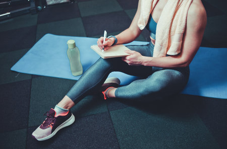 Sporty woman sits on a training mat and writes down future training plans for achieving great results in gymの写真素材