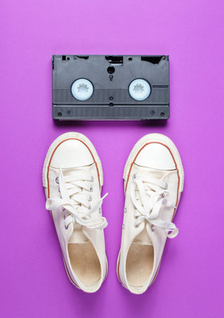 Pop culture attributes 80s. Retro white sneakers shoes, videotape on a purple background. Top viewの写真素材