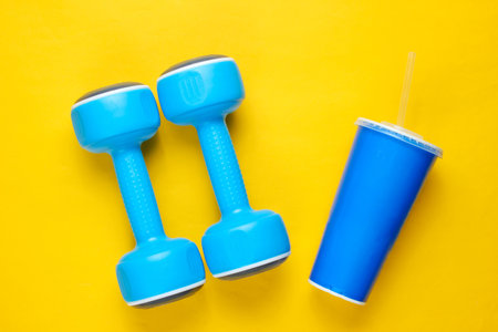 Two blue plastic dumbbells, sport cup on yellow background Sport, fitness concept. Top view. Minimalismの写真素材