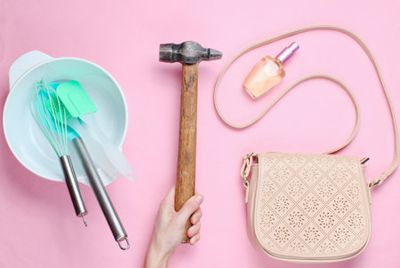 Women choose man's work. Female hand holding hammer on the background of bowl with kitchen tools, bags and bottle of perfume. Pink background, top viewの写真素材