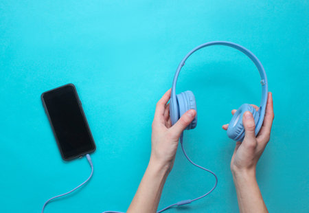 Female hands holding headphones connected to a smartphone on blue background. Top view. Flat lay...