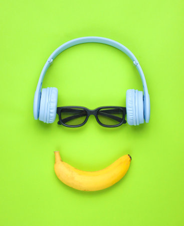Minimalism flat lay concept. Smiling face listens to music Headphones, 3D glasses, banana on a green background.の写真素材