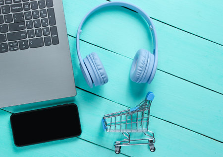 Online shopping. Buyer concept, consumer. Laptop, headphones, smartphone, toy shopping trolley on blue wooden background. Top viewの写真素材