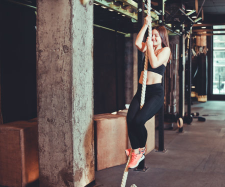 Fit girl climbing up the rope at the gym. Young strong woman performing gymnastics workout. ...