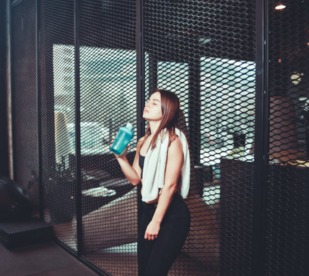 Time for a break. Tired fit girl in sportswear drinks water from  bottle with a towel over her...