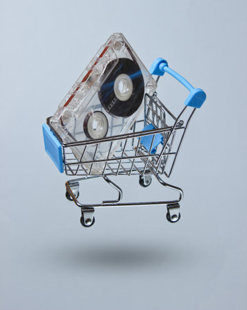 Retro music fan concept.80s. Audio cassette in a toy shopping trolley on a gray background. Photo with shadow, minimalismの写真素材