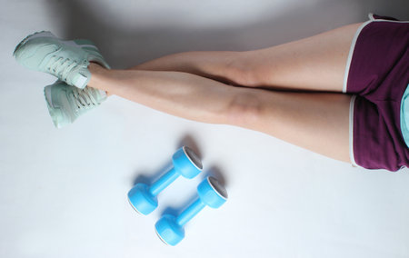 Legs of a young sports woman in shorts and sneakers, plastic dumbbells on a white background. Mimalism fitness concept. Top viewの写真素材