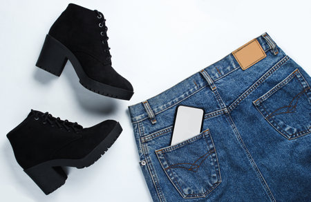 Top view trendy clothes, shoes on a white background. Denim skirt, black boots, smartphone in the back pocketの写真素材