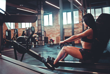 Purposeful fit brunette in sportswear sitting on the simulation rowing exercise machine. Functional...