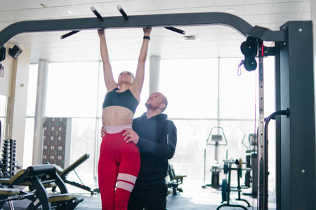 Couple training. Man helps young woman to pull up on the bar in the gymの写真素材