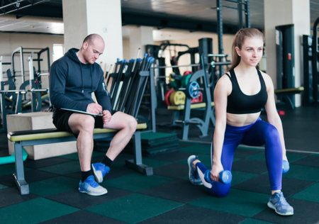Fitness instructor supervises and notes in notebook results of training young athletic blonde performing exercises lunges with dumbbells in her hands in the gymの写真素材