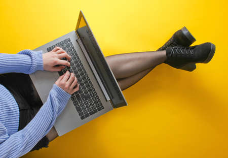 Business woman in shorts, tights and boots is typing on a laptop while sitting on a yellow...