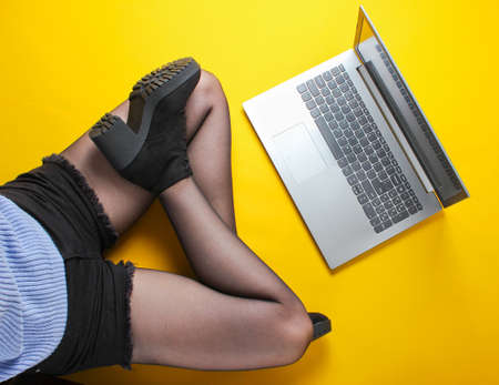 Fashionable woman in shorts, tights and boots use laptop while sitting on yellow background....