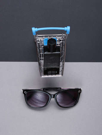 Buyer concept. Perfume bottle in mini shopping trolley, sunglasses on paper background. Top view