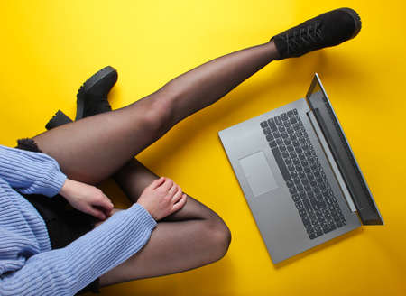 Fashionable woman in shorts, tights and boots use laptop while sitting on yellow background. Freelance, blogger, online worker concept. Top viewの写真素材