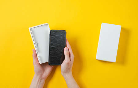 Female hands holding new smartphone in the box. Unboxing Top view on yellow background, minimalism