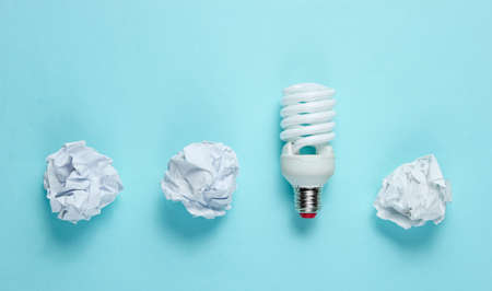 Energy saving light bulb and crumpled paper balls on blue background. Minimalistic business concept,...