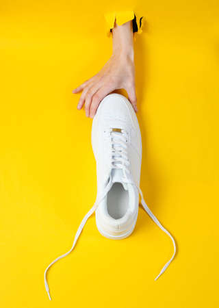 Female hand holds white sneakers through torn yellow paper. Minimalistic creative fashion conceptの写真素材