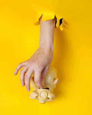 Female hand puts coin into the piggy bank through torn yellow paper background. Minimalistic...