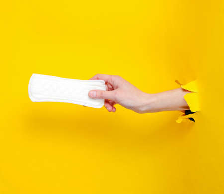 Female hand holds daily sanitary pad through torn yellow paper background. Minimalistic fashion conceptの写真素材