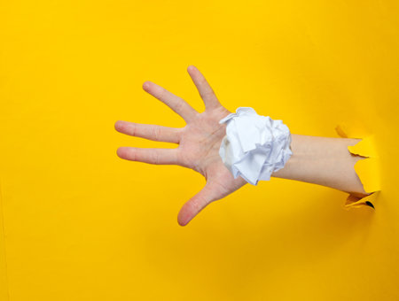 Female hand throws white crumpled ball of paper through the torn yellow paper background. Minimalistic idea conceptの写真素材