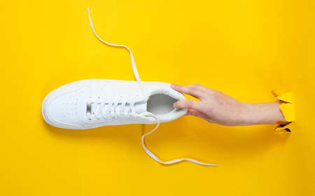 Female hand holds white sneakers through torn yellow paper. Minimalistic creative fashion concept