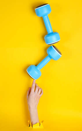 Female hand takes blue plastic dumbbell through torn yellow paper. Minimalistic sport conceptの写真素材