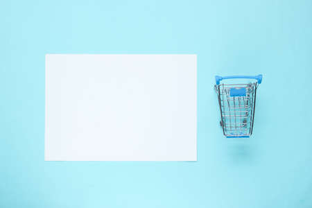 White sheet of paper for copy space, mini shopping trolley on blue background. Creative shopping backgroundの写真素材