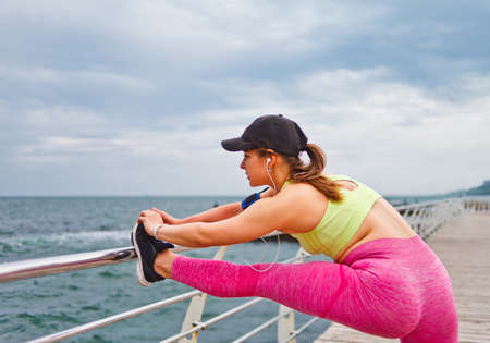 Sports woman in sportswear with headphones in the ears doing stretching leg  exercise before training on the beach outdoorの写真素材